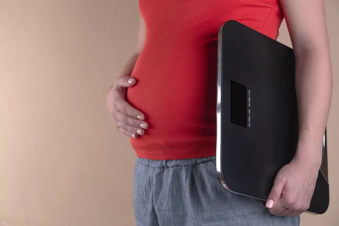 Eine Person in rotem Oberteil und grauer Hose hält eine Digitalwaage in der einen Hand und berührt mit der anderen ihren schwangeren Bauch, was Abnehmen ohne Diät während der Schwangerschaft symbolisiert.
