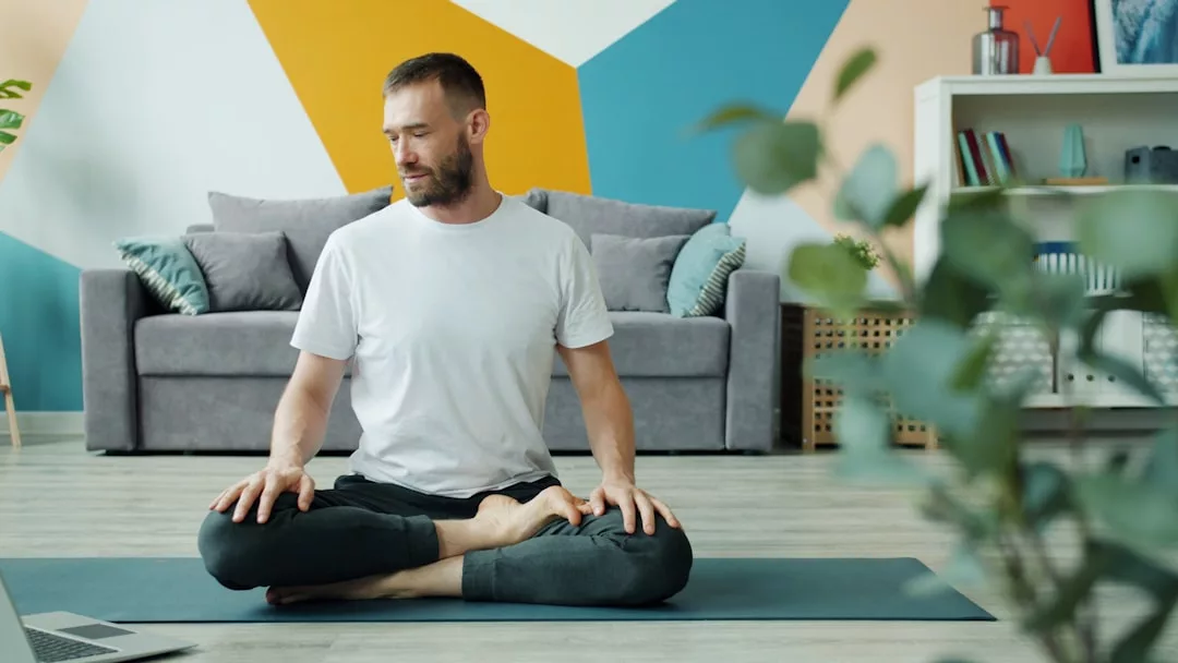 Ein Mann sitzt im Schneidersitz auf einer Yogamatte in einem Haus und übt Achtsamkeit, während er seinen Oberkörper zur Seite dreht und auf einen Laptop schaut. Im Hintergrund sind ein Sofa und ein Bücherregal zu sehen.