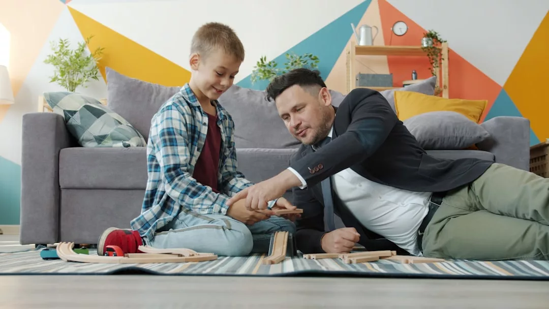 Ein Erwachsener und ein Kind sitzen auf dem Boden eines Wohnzimmers und bauen gemeinsam eine Holzeisenbahn zusammen - eine spielerische Aktivität, die die kindliche Entwicklung unterstützt und die Kreativität fördert.