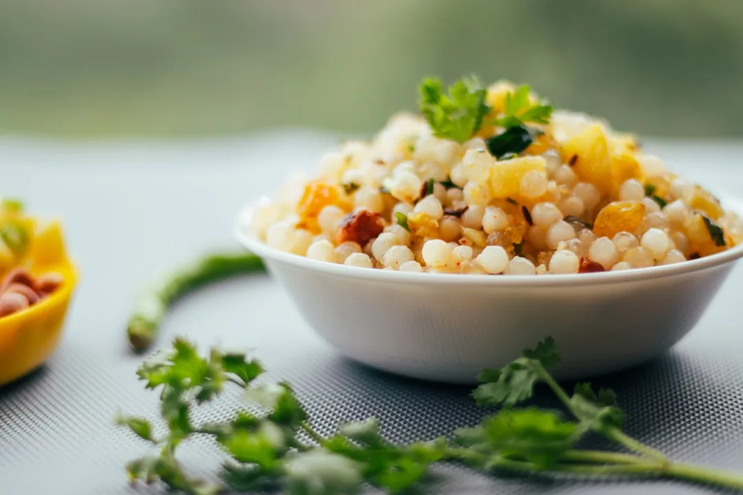 Eine weiße Schüssel mit gekochtem Sabudana Khichdi, garniert mit Koriander, auf einem Tisch mit frischem Koriander und einer grünen Chili in der Nähe - perfekt für alle, die ein schmackhaftes Essen genießen wollen, während sie die Vorteile des Intervallfastens erkunden.