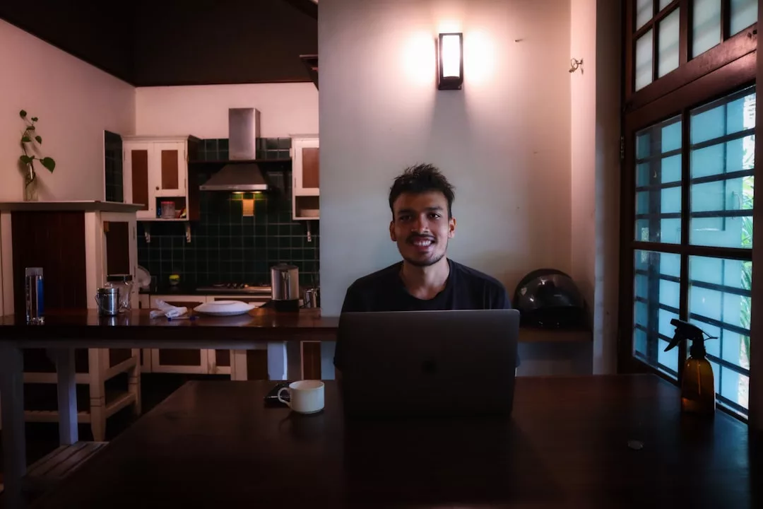 Ein Mann sitzt mit Laptop und Kaffeetasse an einem Tisch in einer modernen Küche und lächelt in die Kamera - das fängt den Geist von Workation Deutschland perfekt ein.