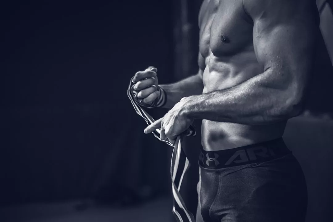 Eine hemdsärmelige, muskulöse Person in Sportkleidung umwickelt ihre Hände mit einer gestreiften Bandage und bereitet sich auf eine Übung oder ein Training vor, wobei sie vielleicht Tipps zur Behandlung von Muskelkater befolgt.