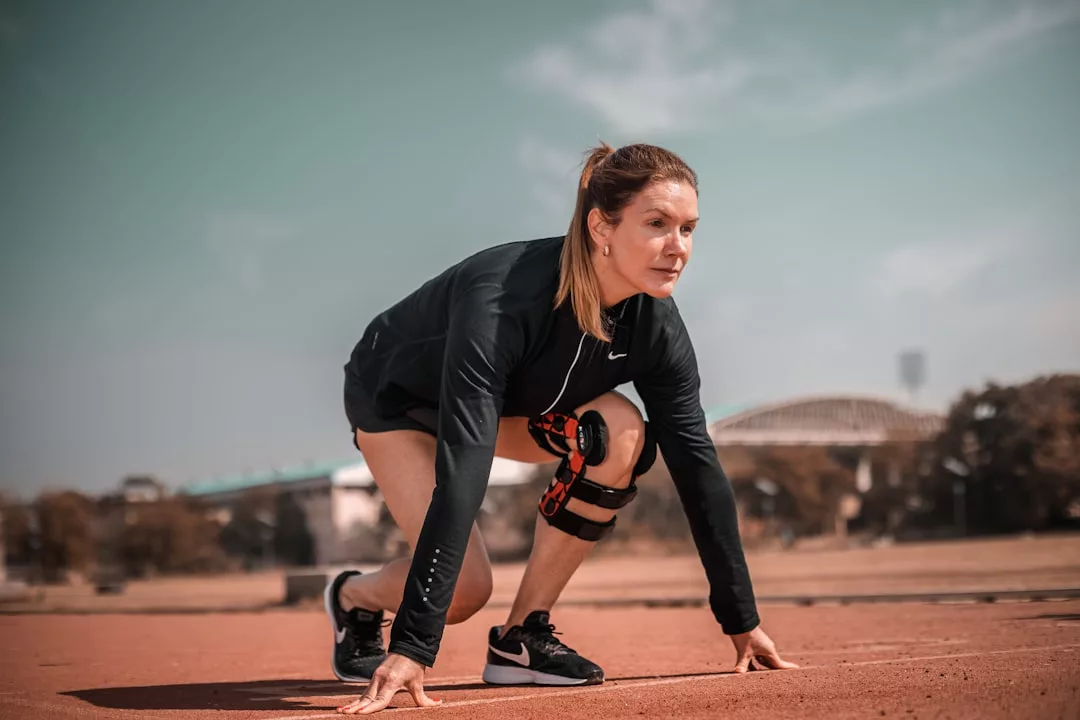Eine Frau in Sportkleidung und mit Knieschiene kniet an der Startlinie einer Outdoor-Laufbahn und bereitet sich auf den Sprint vor - bereit, Sportverletzungen mit gezielter Behandlung und Tipps zu überwinden.