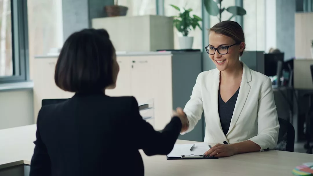 Zwei Frauen sitzen in einem Büro an einem Schreibtisch und schütteln sich die Hände - ein perfektes Beispiel für Netzwerken und berufliche Kontakte knüpfen. Die eine trägt einen weißen Blazer und eine Brille, die andere hat kurze dunkle Haare und eine dunkle Jacke.