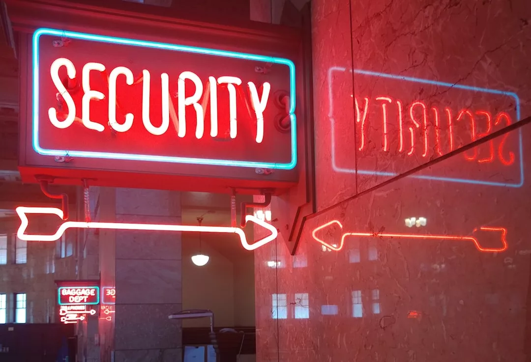 Eine Leuchtreklame mit der Aufschrift "SECURITY" und einem roten Pfeil weist nach rechts und spiegelt sich in einer Marmorwand; im Hintergrund leuchtet eine Leuchtreklame mit der Aufschrift "BAGGAGE DEPT", die ein Gefühl von beruflicher Sicherheit an diesem modernen Arbeitsplatz hervorruft.