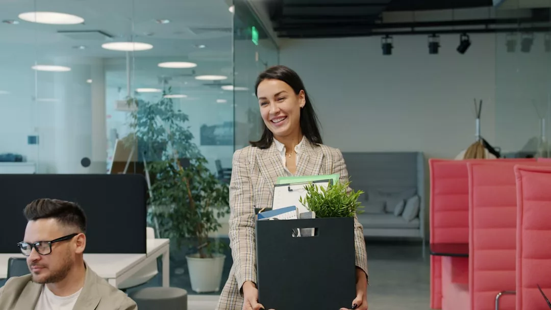 Eine Frau steht in einem Büro und hält eine Schachtel mit persönlichen Gegenständen und einer Pflanze in der Hand, die eine berufliche Veränderung symbolisiert, während ein Mann an einem Schreibtisch in der Nähe sitzt und arbeitet.