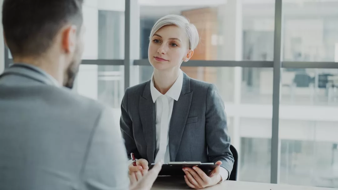 Eine Frau in einem grauen Anzug, die ein Klemmbrett in der Hand hält, hört einem Mann während einer beruflichen Besprechung zu, als sie wertvolle Tipps für den nächsten Karriereschritt in einem modernen Büro besprechen.
