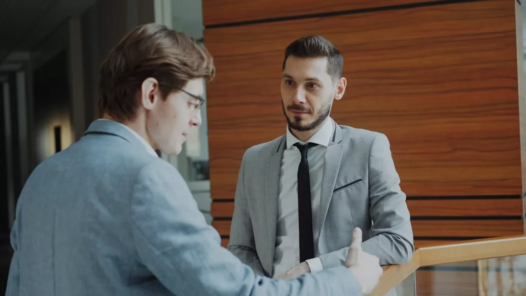 Zwei Männer in Businesskleidung unterhalten sich drinnen an einem Holzgeländer - einer spricht, während der andere aufmerksam zuhört und wertvolle Karrieretipps für eine erfolgreiche Karriere austauscht.