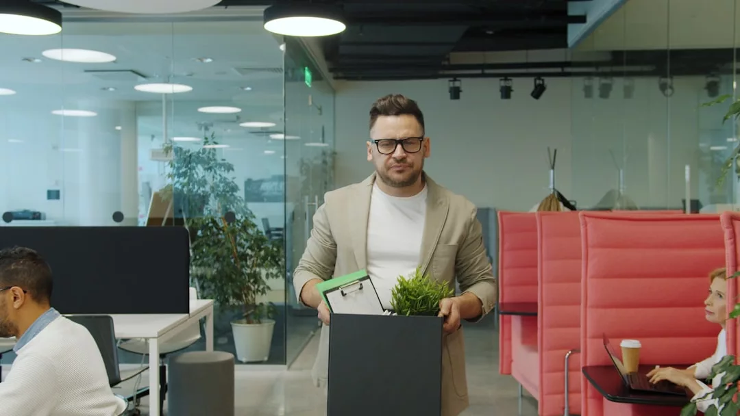 Ein Mann über 40 hält eine Box mit Pflanze und persönlichen Sachen und steht für seinen beruflichen Neustart im Büro, während andere arbeiten und eine Frau mit Kaffeetasse sitzt.
