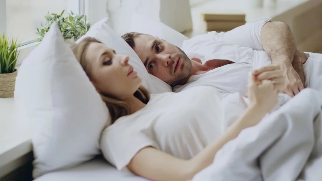 Ein Mann und eine Frau liegen Seite an Seite im Bett. Die Frau scheint mit geschlossenen Augen zu schlafen, während der Mann wach ist und nach vorne schaut. Beide sind weiß gekleidet, was entweder auf Beziehungskonflikte oder eine harmonische Partnerschaft hindeutet.
