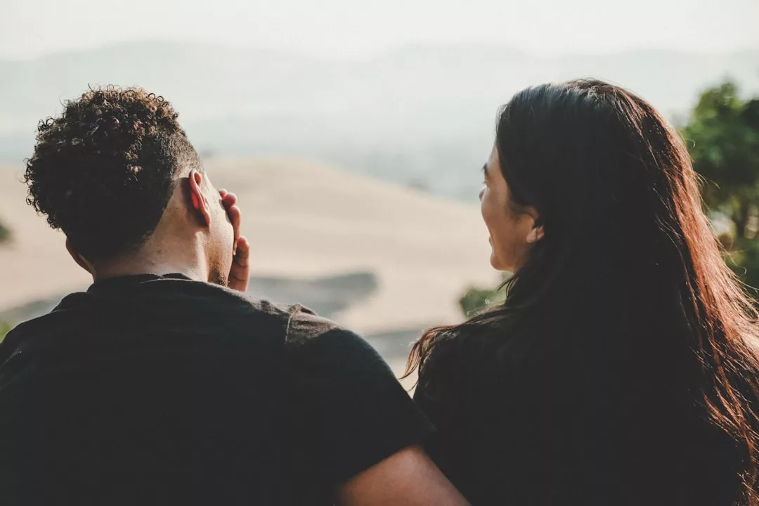 Zwei Menschen sitzen im Freien dicht beieinander und blicken auf eine ferne Landschaft, während der eine den anderen anlächelt - ein Moment, der zeigt, wie Paare gemeinsame Beziehungskrisen erfolgreich meistern können.