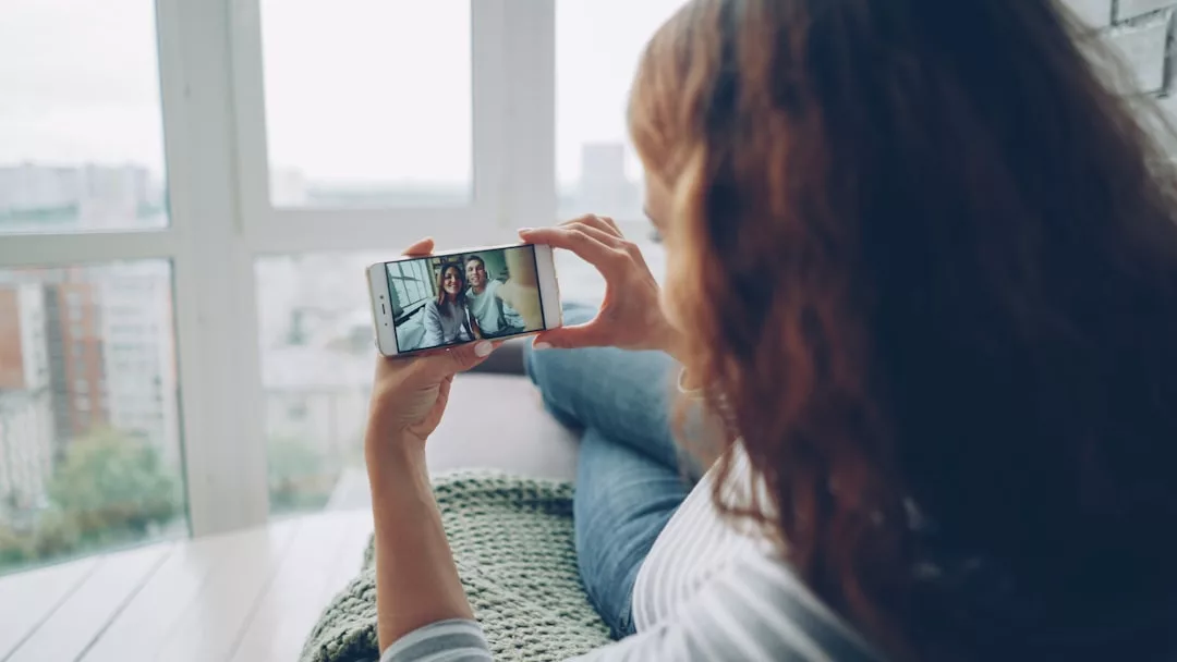 Eine Frau sitzt am Fenster, hält ein Smartphone und macht ein Selfie mit einer anderen Person, die auf dem Bildschirm erscheint - ein schönes Beispiel für Beziehungspflege im Alltag.