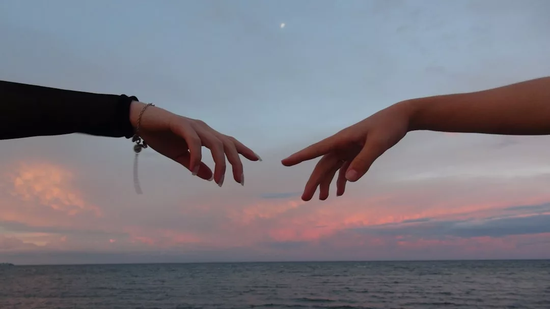 Zwei Hände, die sich einander entgegenstrecken, wobei sich die Finger fast berühren, vor dem Hintergrund des Ozeans und des rosafarbenen Himmels bei Sonnenuntergang, der an die Schönheit der Beziehungen und die stille Poesie der Beziehungsrituale erinnert.