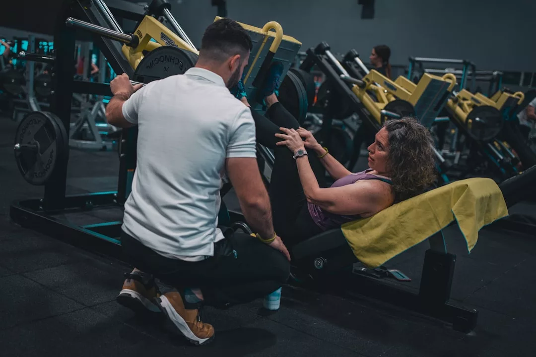 Eine Frau benutzt eine Beinpresse im Fitnessstudio, während ein Trainer in einem weißen Hemd neben ihr kniet und sie beim Beziehungstraining anleitet, um ihre Partnerschaft zu verbessern.