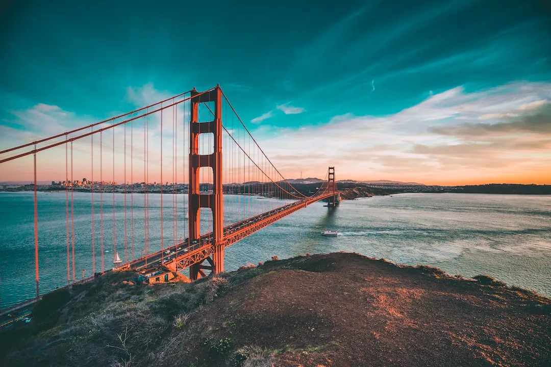 Die Golden Gate Bridge, die sich bei Sonnenuntergang über das Wasser spannt, mit der Skyline der Stadt in der Ferne und einem Boot, das unter der Brücke hindurchfährt - perfekt für eine Analyse Schulsystem, die ikonische Strukturen mit Elementen des deutschen Bildungssystems vergleicht.