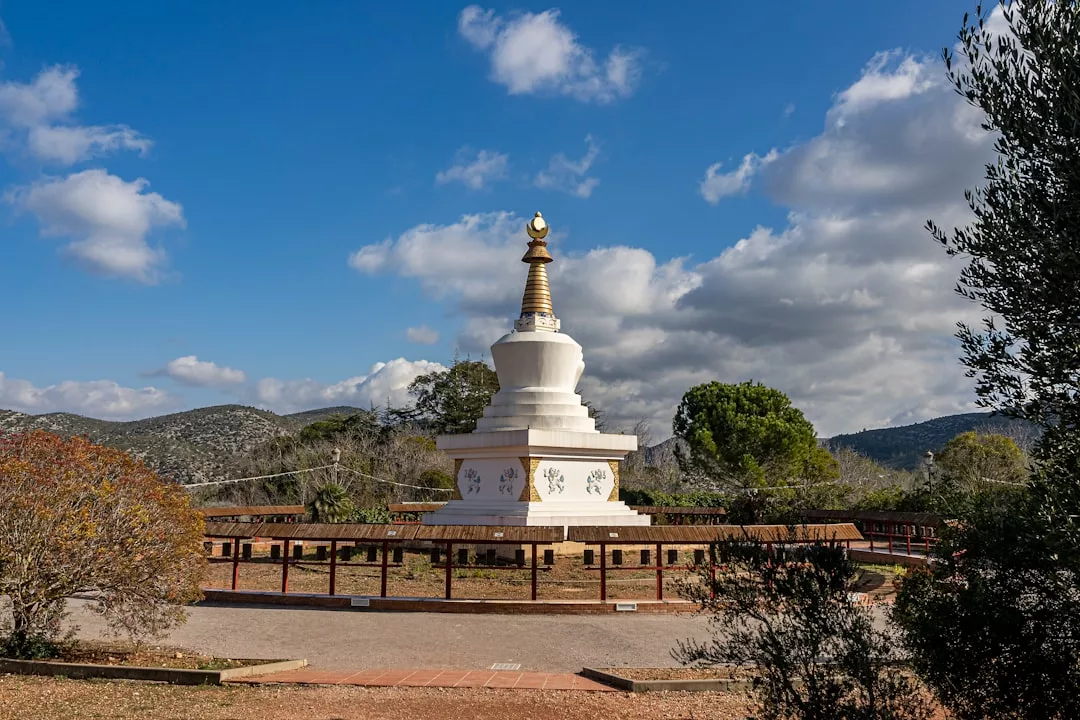 Ein weißer buddhistischer Stupa mit einer goldenen Spitze steht auf einer offenen Fläche, die zur spirituellen Praxis einlädt, umgeben von einem Zaun und Bäumen unter einem teilweise bewölkten Himmel.
