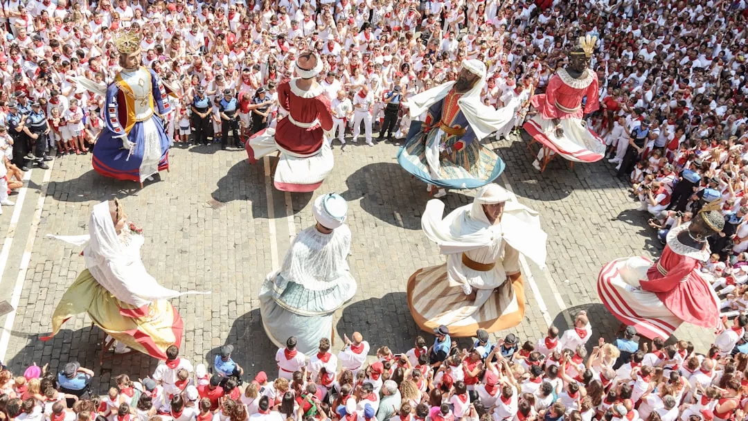 Große kostümierte Figuren ziehen und tanzen in einer belebten Straße, die den Einfluss der Kultur widerspiegelt, während sich die Zuschauer - viele in weißer Kleidung mit roten Schals - versammeln, um dieses lebendige gesellschaftliche Ereignis zu feiern.
