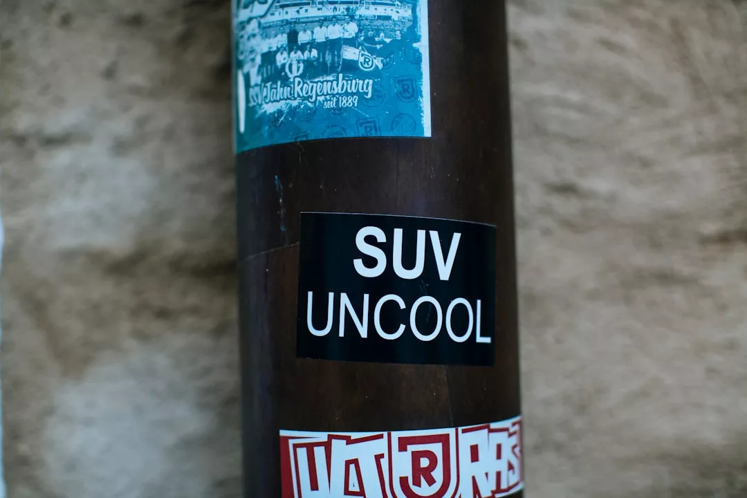 Nahaufnahme eines schwarzen Aufklebers mit der Aufschrift "SUV UNCOOL" auf einem braunen Mast, der den Einfluss auf das städtische Klima hervorhebt, mit anderen Aufklebern und einer strukturierten Wand im Hintergrund.