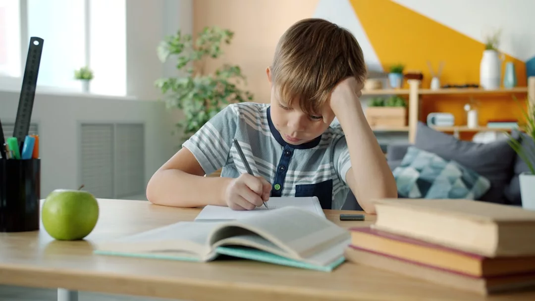 Ein Junge sitzt an einem Schreibtisch und schreibt in ein Notizbuch, den Kopf auf eine Hand gestützt - eine Momentaufnahme der persönlichen Entwicklung und des ruhigen Fokus des Lernprozesses. Ein aufgeschlagenes Buch, ein grüner Apfel und ein Stapel Bücher liegen auf dem Schreibtisch.