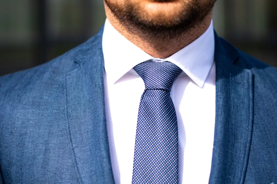 Nahaufnahme eines nach dem Dresscode Bewerbungsgespräch gekleideten Mannes, der einen blauen Anzug, ein weißes Hemd und eine gemusterte blaue Krawatte trägt. Nur die untere Hälfte seines Gesichts und sein Oberkörper sind zu sehen.