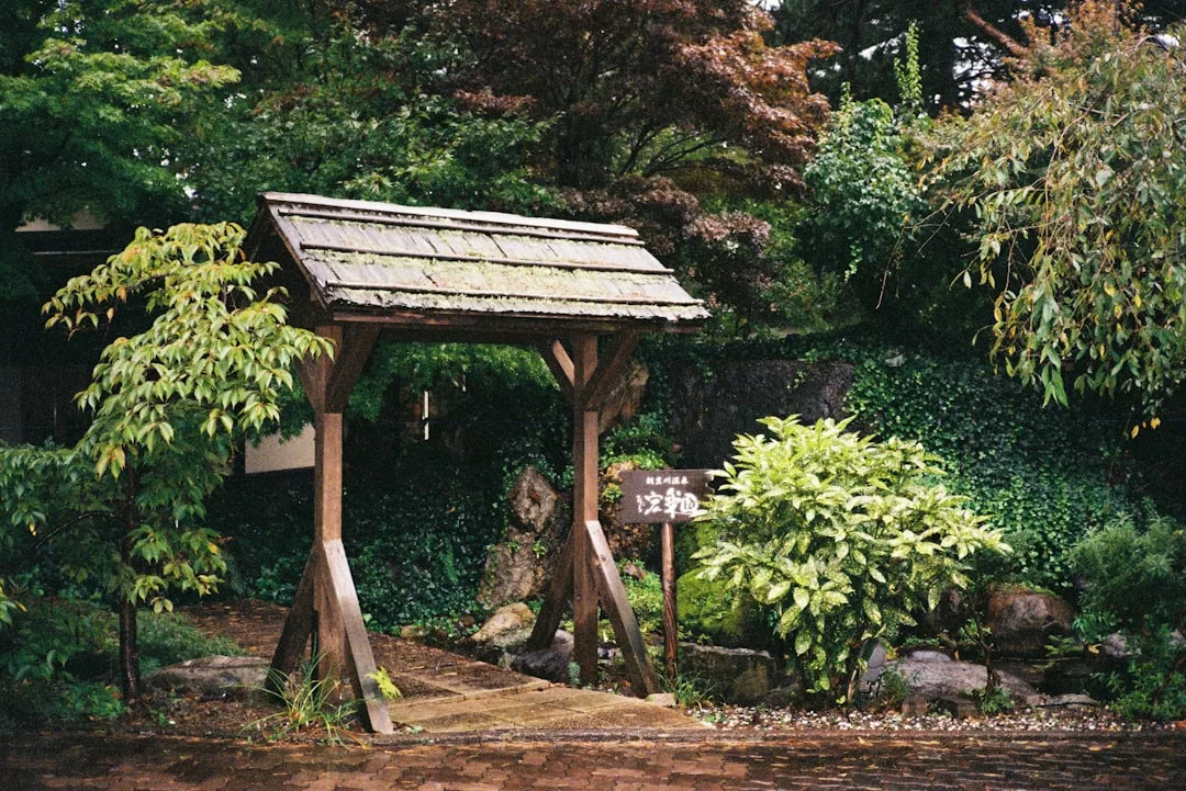 Ein hölzernes Tor steht über einem steinernen Weg, umgeben von grünen Pflanzen und Bäumen, mit einem Schild in japanischer Sprache auf der rechten Seite - eine inspirierende Szene für jedes Gartenjahr oder diejenigen, die neue Gartentipps suchen.