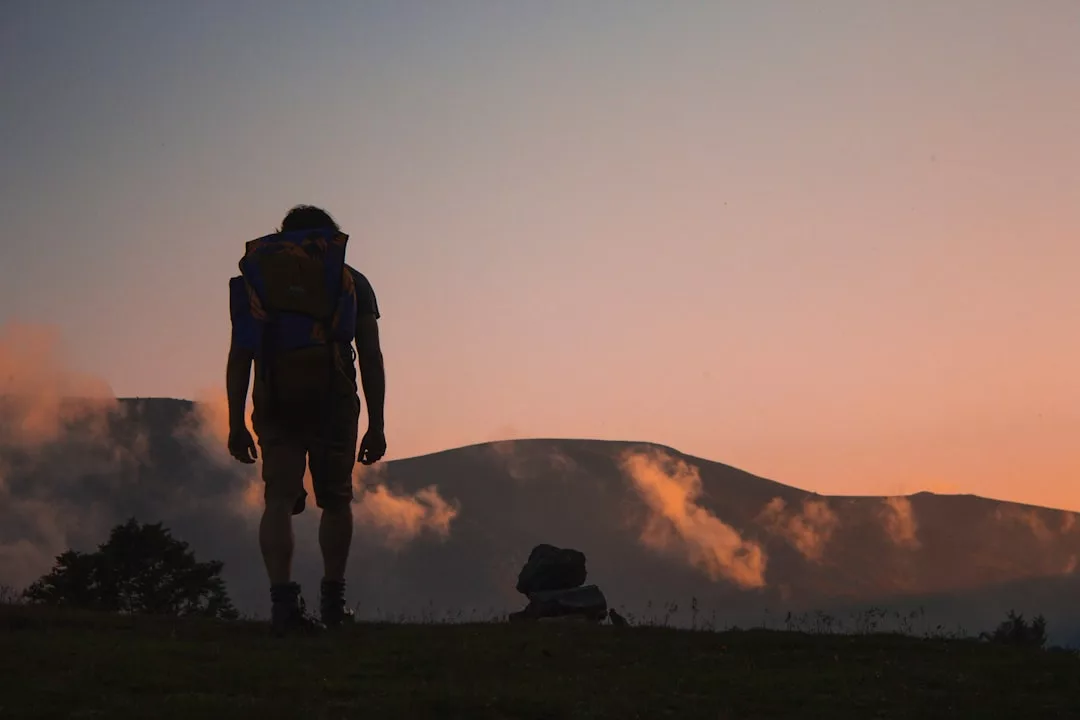 In goldenes Abendlicht getaucht, steht eine Person mit einem Rucksack auf einem grasbewachsenen Hügel, Berge und Wolken im Hintergrund - eine Szene voller Inspiration und Abenteuerlust.