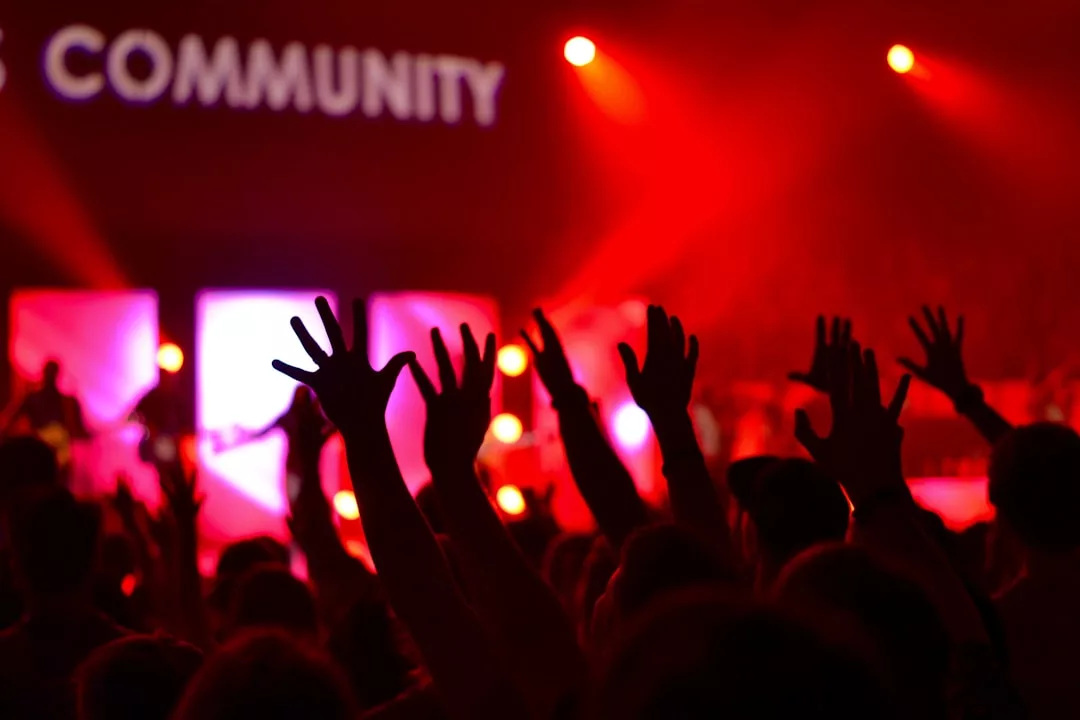 Eine Menschenmenge mit erhobenen Händen bei einer Indoor-Veranstaltung, beleuchtet von rotem Bühnenlicht, mit dem Wort "COMMUNITY" im Hintergrund - eine Atmosphäre, die an eine Musiktherapie-Anwendung erinnert, die Menschen zusammenbringt.