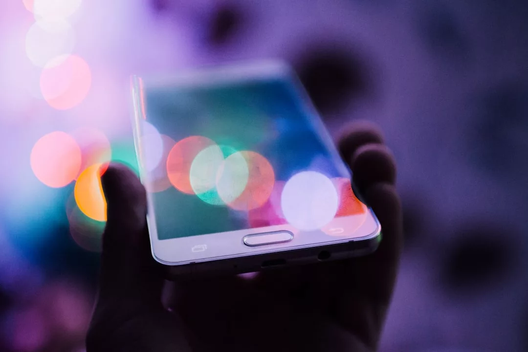 Eine Hand hält ein weißes Smartphone, auf dessen Bildschirm sich bunte, verschwommene Lichter spiegeln und die Auswirkungen der Digitalisierung auf die moderne Arbeitswelt symbolisieren.