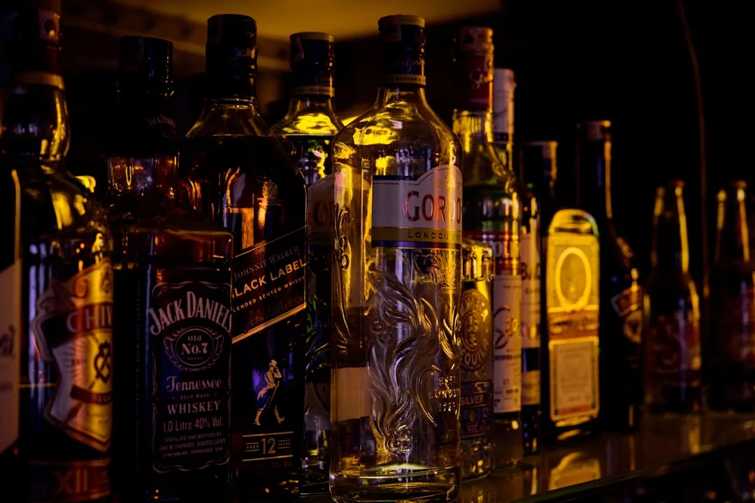 In einem schwach beleuchteten Barregal stehen verschiedene Spirituosenflaschen, darunter Whiskey, Wodka und Gin, die sowohl die Faszination als auch die Risiken von Alkohol für die Gesundheit verdeutlichen.