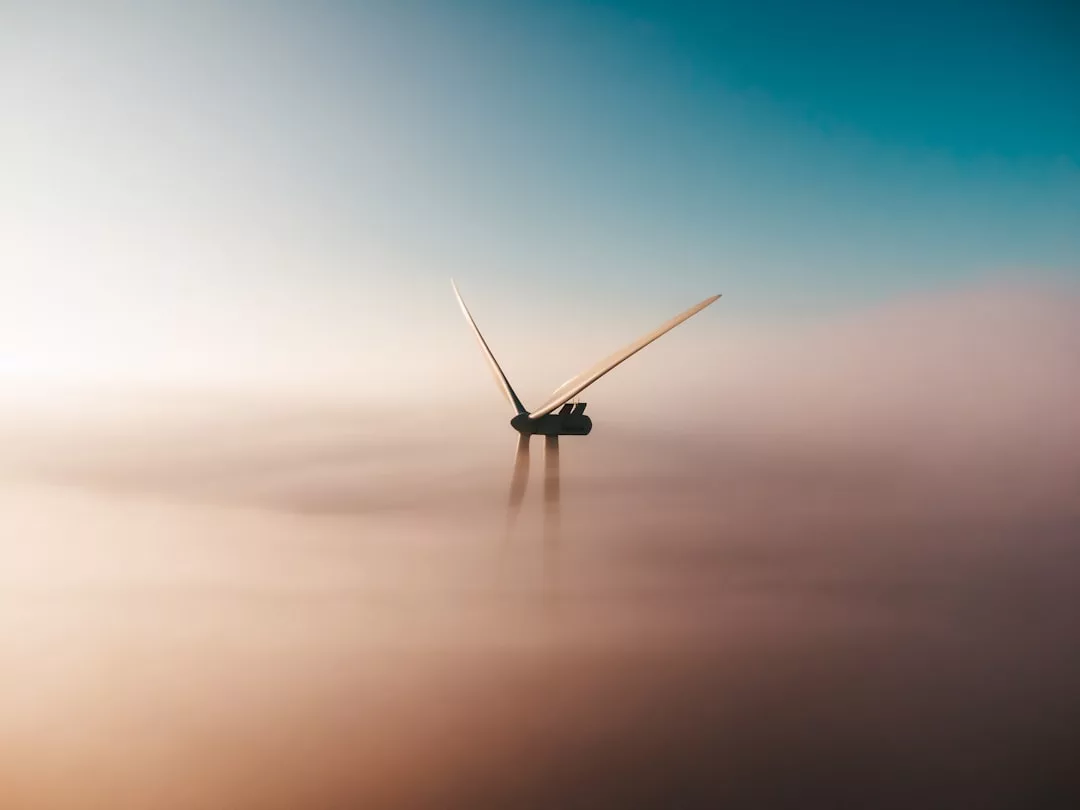 Eine Windturbine erhebt sich über einer Nebelschicht, ihre Flügel sind ausgefahren und nutzen die Windenergie vor einem Himmel, der von Blau zu warmen Tönen wechselt, um die positiven Auswirkungen auf unsere Umwelt hervorzuheben.