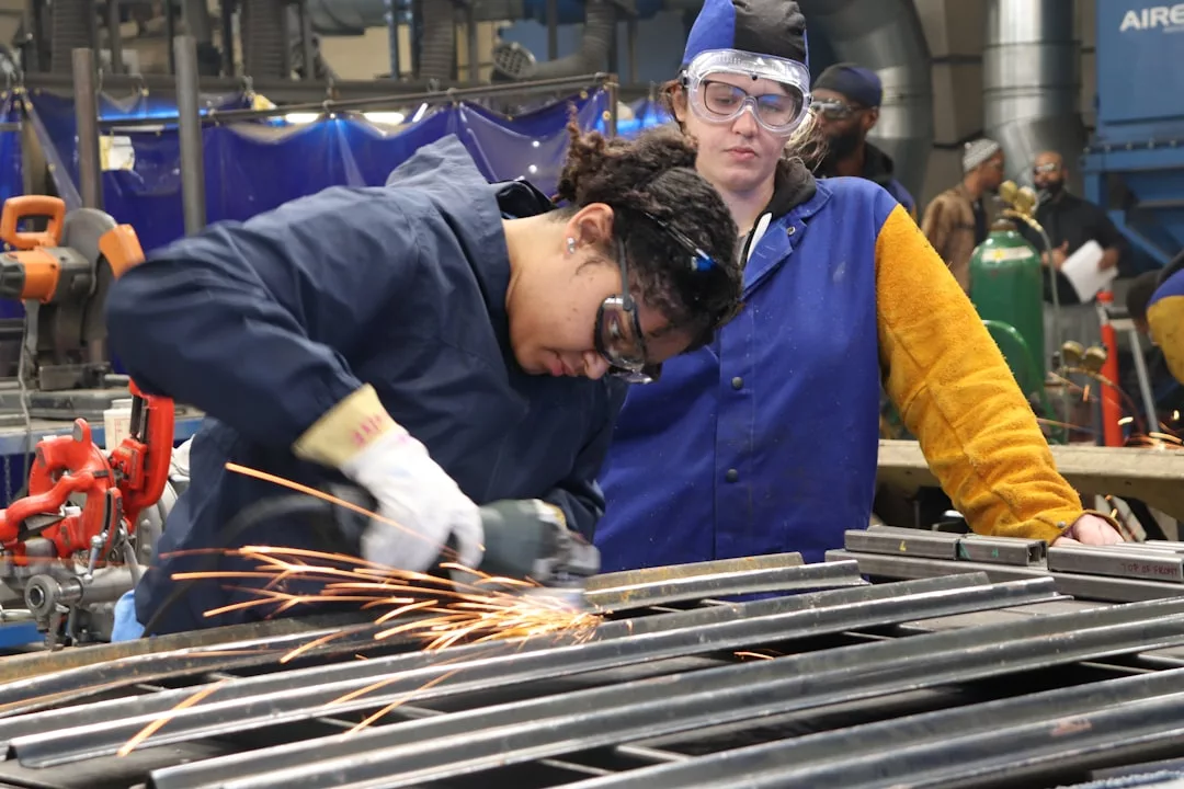 Zwei Personen in Schutzkleidung arbeiten in einer Metallwerkstatt; die eine bedient eine Schleifmaschine, die Funken erzeugt, während die andere zuschaut, was die Bedeutung von Berufsausbildung in der modernen Arbeitswelt unterstreicht.