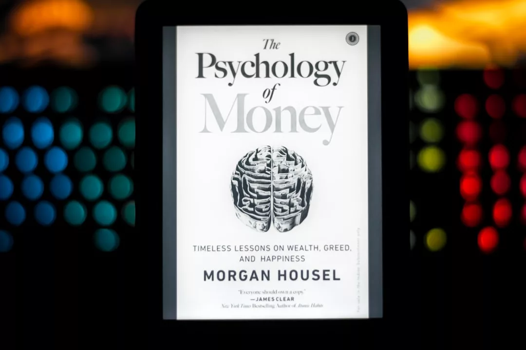 Ein digitales Tablet zeigt das Cover von "The Psychology of Money" von Morgan Housel, das eine Münze in Form eines Gehirns und schwarzen Text auf weißem Hintergrund zeigt - ein aufschlussreicher Einblick in die Börsenpsychologie und die Bedeutung von Emotionen an der Börse.