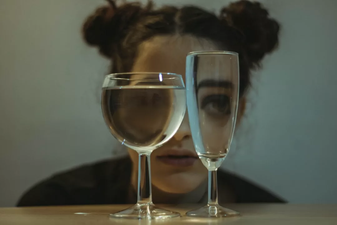 Eine Person mit zwei Dutts im Haar blickt durch ein Weinglas und eine mit Wasser gefüllte Sektflöte, wobei ihre Gesichtszüge verzerrt sind - ein Hinweis darauf, wie emotionale Intelligenz unsere Wahrnehmung und Interpretation anderer Menschen verändern kann.
