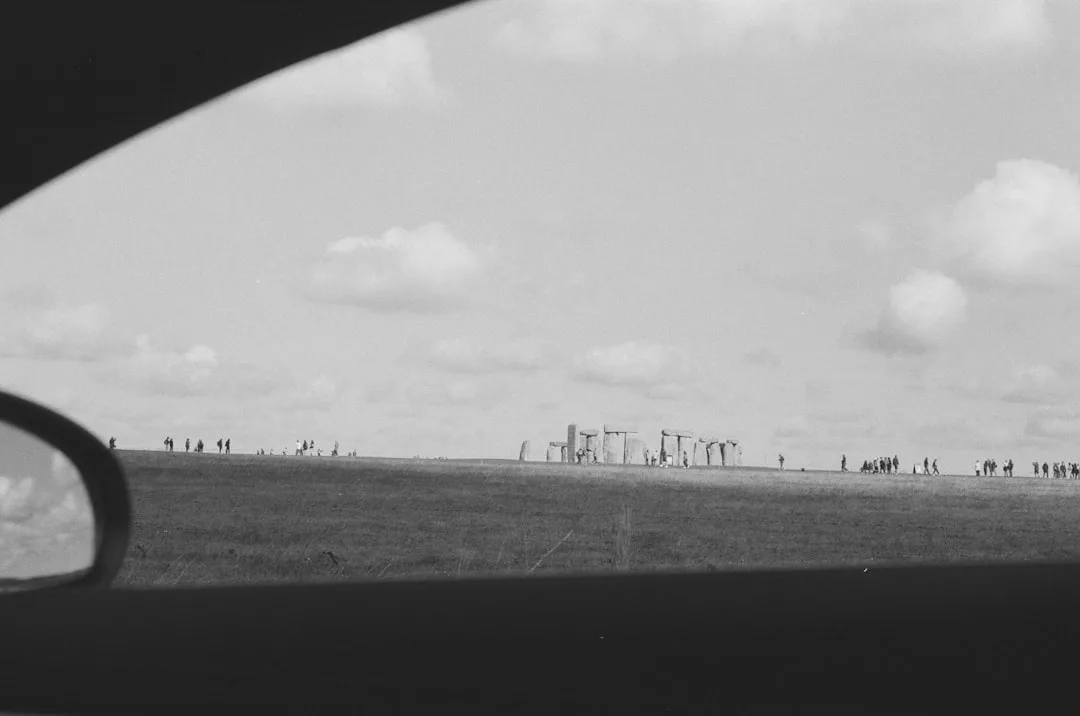 Schwarz-Weiß-Foto von Stonehenge durch ein Autofenster gesehen, mit Menschen, die in der Ferne unter einem teilweise bewölkten Himmel spazieren gehen - eine zeitlose Szene, die an die sich entwickelnde Energiepolitik in Deutschland erinnert.