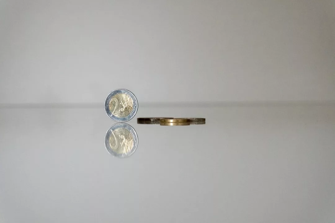 Eine Zwei-Euro-Münze steht aufrecht auf ihrem Rand neben zwei anderen, die flach auf einer spiegelnden Oberfläche liegen, wodurch ein Spiegeleffekt entsteht, der zum Nachdenken über die Bedeutung von Geld in unserer Gesellschaft einlädt.