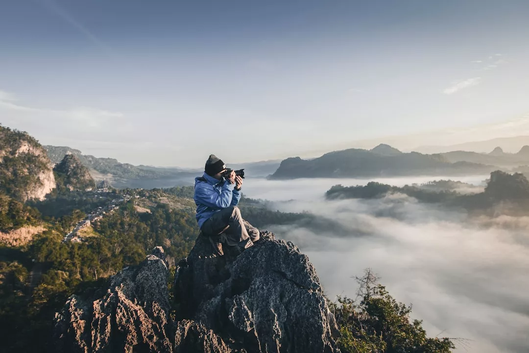 Eine Person mit blauer Jacke und Mütze sitzt auf einem felsigen Gipfel und fotografiert ein nebelverhangenes Tal und ferne Berge bei Sonnenaufgang und findet Bedeutung in der atemberaubenden Szene.