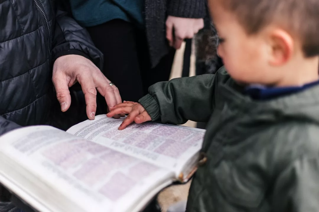 Ein Kind berührt ein aufgeschlagenes Buch, möglicherweise eine Bibel, während ein Erwachsener auf eine Stelle auf der Seite zeigt - eine alltägliche Szene, die die Bedeutung von Kindererziehung unterstreicht. Im Hintergrund sind weitere Personen zu sehen.