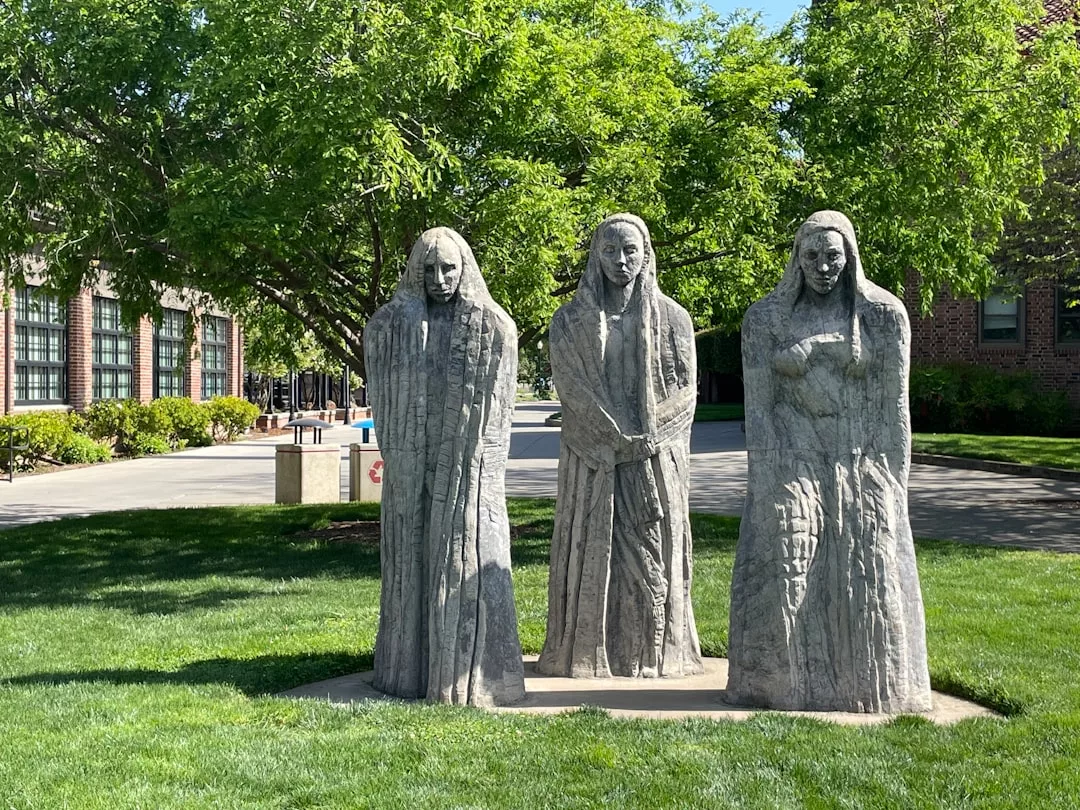 Drei steinerne Statuen von stehenden Frauen in langen Gewändern stehen im Freien auf einem Betonsockel, umgeben von Gras und Bäumen - ein beeindruckendes Beispiel für Kunst mit tiefgründiger Bedeutung.