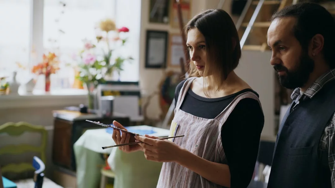 Zwei Personen stehen in einem Raum, eine mit Pinsel und Palette in der Hand, umgeben von Kunstzubehör und Blumen, die die Bedeutung von Kunstpädagogik in der Schule widerspiegeln.
