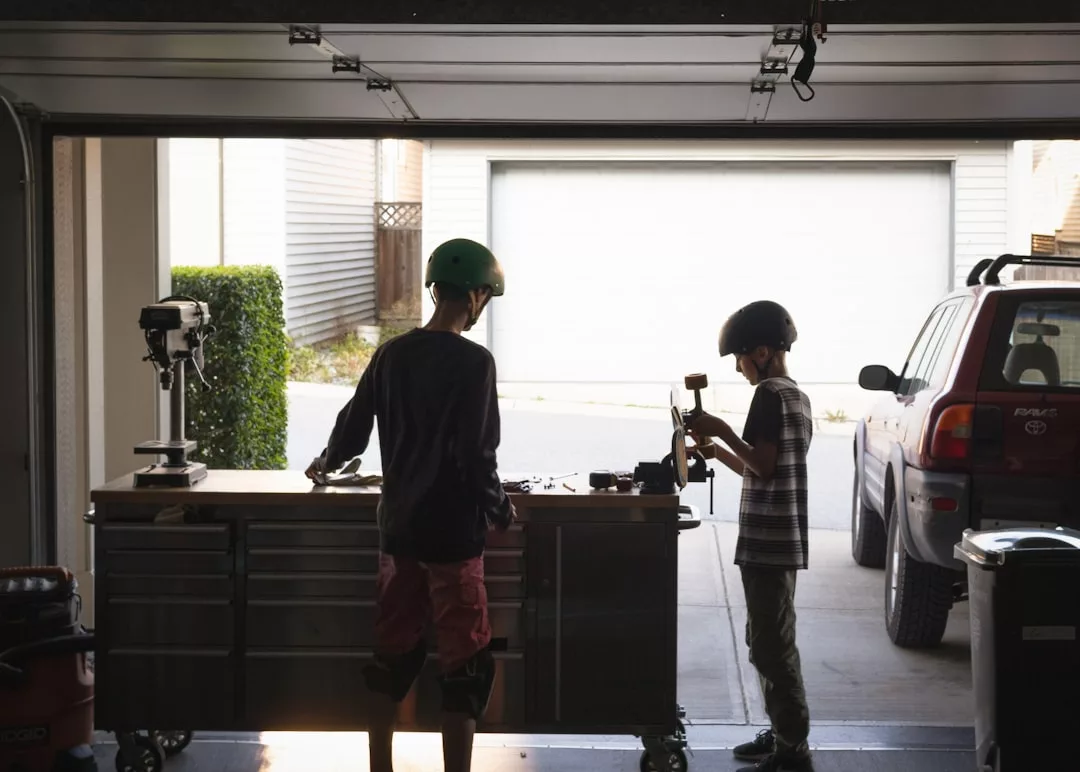 Zwei Kinder mit Helmen und Schützern arbeiten gemeinsam an einem Projekt in einer Garagenwerkstatt und zeigen die Bedeutung von Freundschaften, während sie in der Nähe eines geparkten Geländewagens und eines offenen Garagentors zusammenarbeiten.