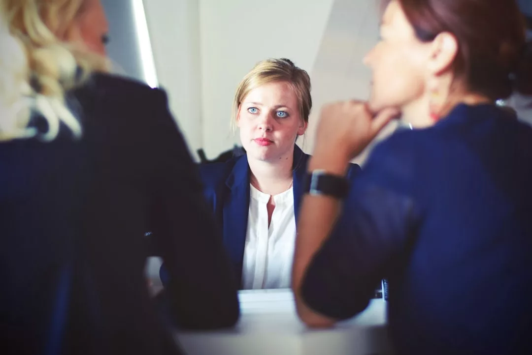 Drei Frauen in Business-Kleidung sitzen an einem Tisch und diskutieren über Arbeitsrecht. Eine Frau ist der Kamera zugewandt, während die beiden anderen Frauen aus den Unternehmen von hinten und im Profil zu sehen sind.