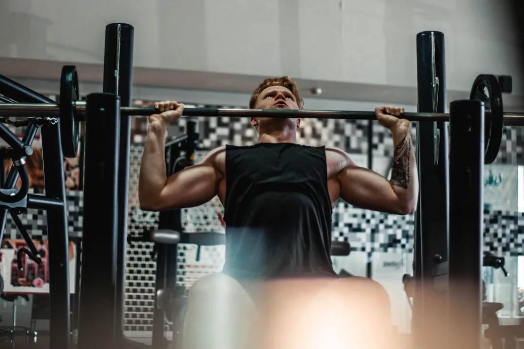 Eine Person in einem schwarzen Tank-Top führt eine Überkopf-Hantelpresse aus, während sie in einem Fitnessstudio sitzt, und zeigt damit ihre Hingabe zu körperlicher Fitness und Krafttraining.