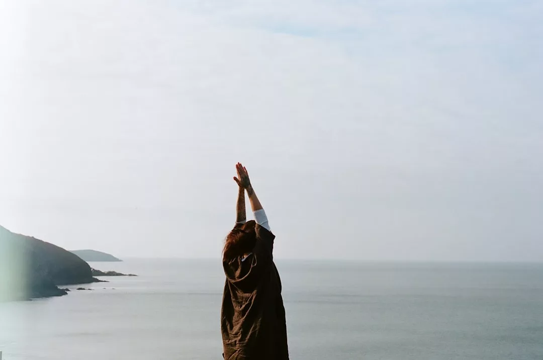 Eine Person, die mit erhobenen Armen in Küstennähe steht und unter einem bewölkten Himmel auf das Meer blickt - ein stiller Moment, der die Bedeutung von Achtsamkeit einfängt. Im Hintergrund sind die felsige Küste und das ferne Land zu sehen.