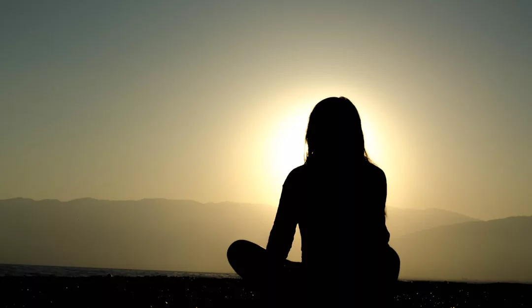 Silhouette einer Person, die im Schneidersitz im Freien sitzt und Achtsamkeit übt, während die Sonne hinter ihr unter- oder aufgeht, mit Bergen im Hintergrund - ein friedlicher Moment inmitten der modernen Welt.
