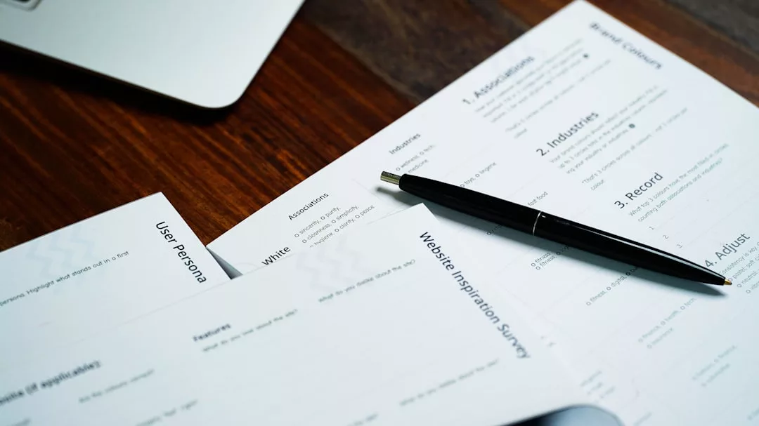 Ein Stift ruht auf gedruckten Dokumenten mit der Aufschrift "Website Inspiration Survey" und "User Persona", die die Bedeutung von Arbeitgeberbewertung in der modernen Arbeitswelt hervorheben, während ein Laptop teilweise auf dem Holztisch steht.