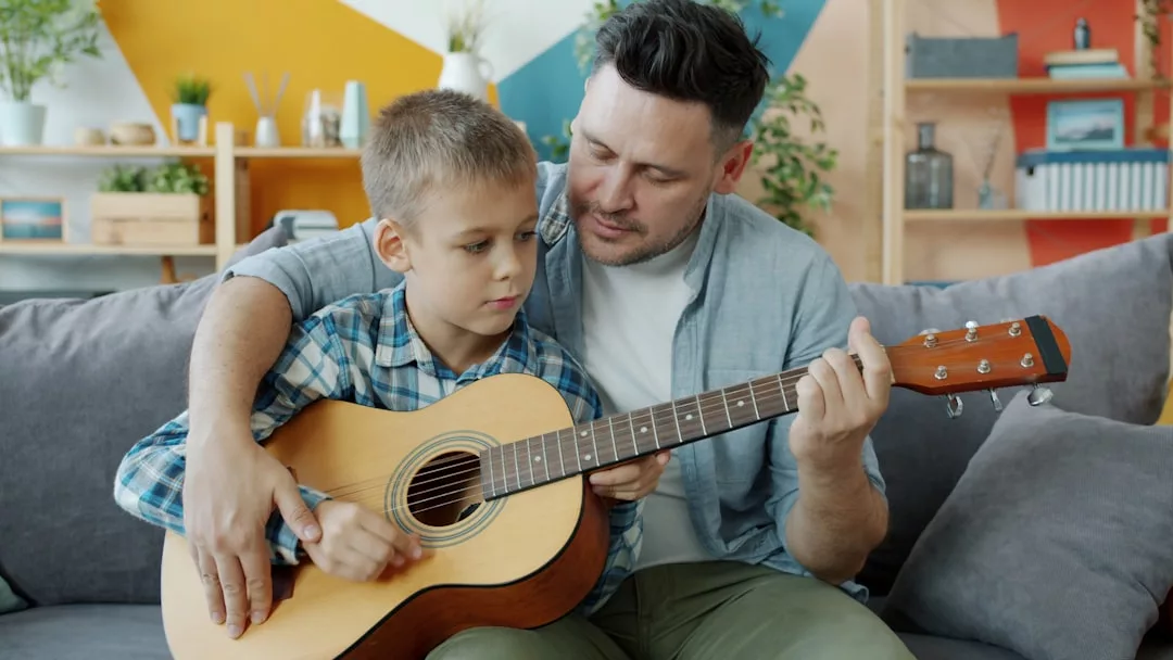 Ein Erwachsener hilft einem Jungen beim Spielen einer Akustikgitarre, während sie gemeinsam auf einer Couch in einem hell dekorierten Wohnzimmer sitzen, um die Bildungsqualität und die Verbindung in der Gesellschaft zu fördern.