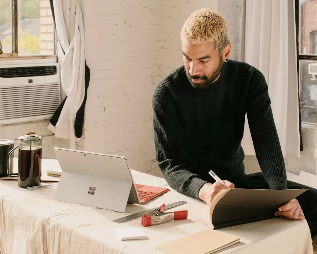 Eine Person sitzt an einem Schreibtisch mit einem Laptop, einem Notebook, einer Kaffeemaschine und Büromaterialien und untersucht die Bedeutung der Bildungstechnologie in der modernen Gesellschaft, während sie in ein Notebook schreibt und auf den Laptop-Bildschirm schaut.