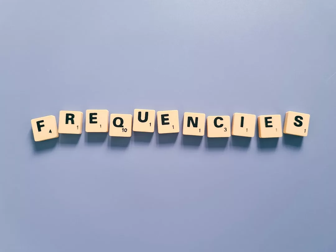 Scrabble-Kacheln aus Holz buchstabieren das Wort "FREQUENCIES" auf einer schlichten, hellblauen Oberfläche und erinnern an Themen der Elektronik und den Begriff Frequenz.