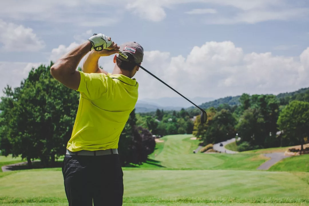 Ein Golfer in einem gelben Hemd schwingt seinen Schläger an einem sonnigen Tag, mit Blick auf einen modernen grünen Golfplatz mit Bäumen und Hügeln im Hintergrund.