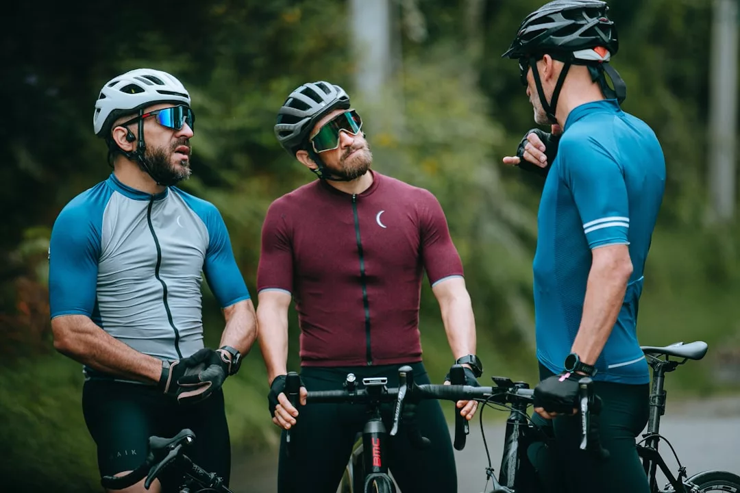 Drei Radfahrer mit Helmen, Sonnenbrillen und hochwertiger Sportbekleidung stehen draußen mit ihren Fahrrädern und unterhalten sich.
